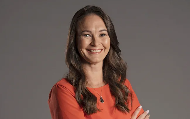 A woman with long hair wearing a red top smiles, arms crossed. Gray background.