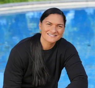 A person with long black hair smiles, sitting by a blue pool, wearing a black sweater.