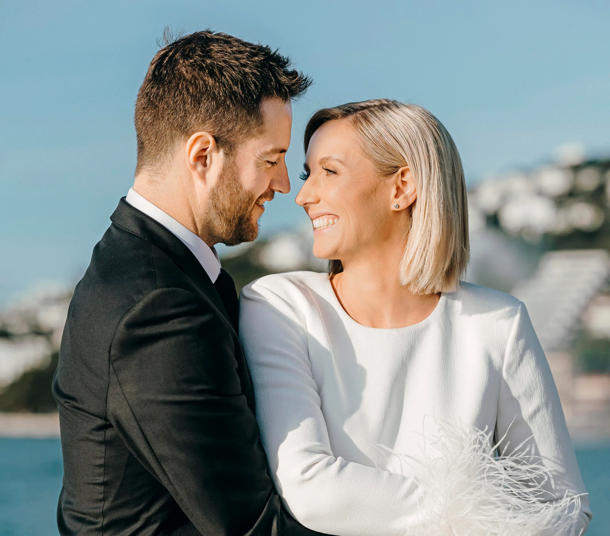 A couple in formal attire smiling at each other with a blurry seaside background.