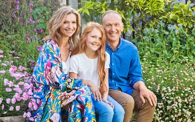 Family of three sitting together in a lush garden with colorful flowers, smiling at the camera.