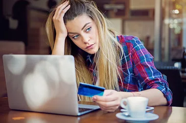 Young woman with long hair looks frustrated, sitting at a table with a laptop, holding a credit card, and drinking coffee.