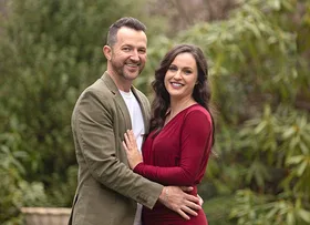Couple smiling and embracing in a garden, with a man in a green jacket and a woman in a red dress.
