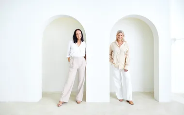 Two women smiling and standing in arched doorways, wearing light-colored outfits in a minimalistic white room.