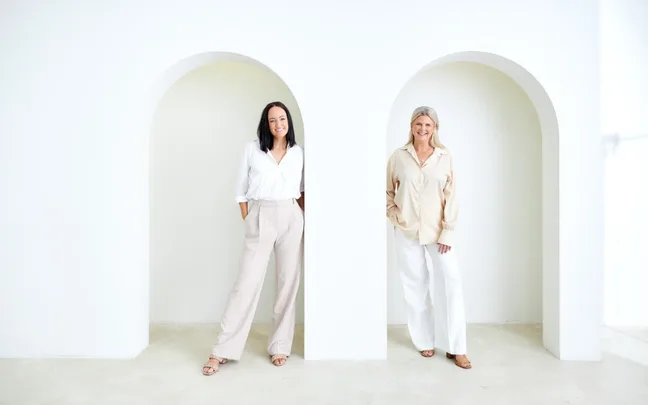 Two women smiling and standing in arched doorways, wearing light-colored outfits in a minimalistic white room.
