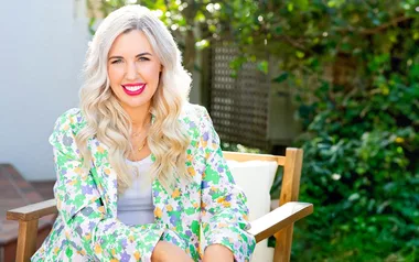 A woman with long blonde hair in a floral blazer sits outside on a wooden chair, smiling brightly.