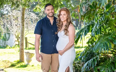 Couple posing outdoors, woman in white dress tenderly holding her belly, surrounded by trees and greenery.