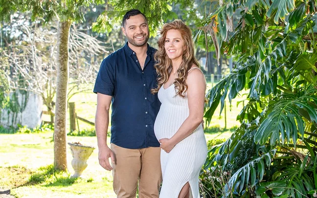 Couple posing outdoors, woman in white dress tenderly holding her belly, surrounded by trees and greenery.