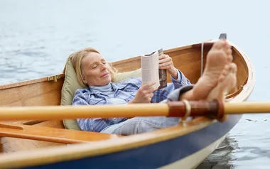 Woman relaxing in a small boat on a lake, reading a book with her feet propped up.