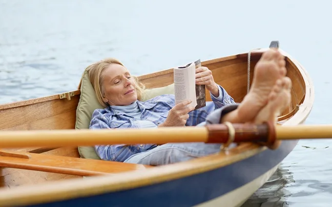 Woman relaxing in a small boat on a lake, reading a book with her feet propped up.