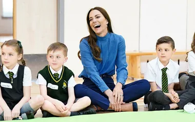 A woman in a blue blouse sits cross-legged, laughing with children in uniform around her.