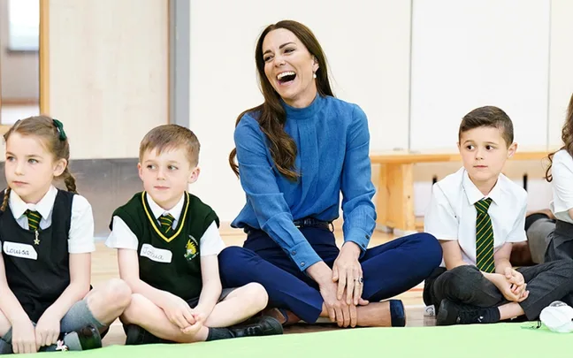 A woman in a blue blouse sits cross-legged, laughing with children in uniform around her.