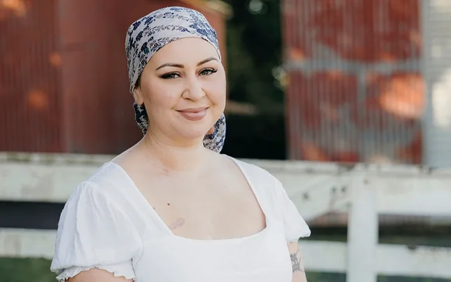 Female smiling, wearing a patterned headscarf and white top, standing outdoors with a barn in the background.