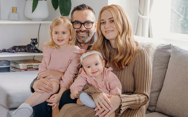 A family of four smiling on a sofa, with parents and two young children in light pink outfits.