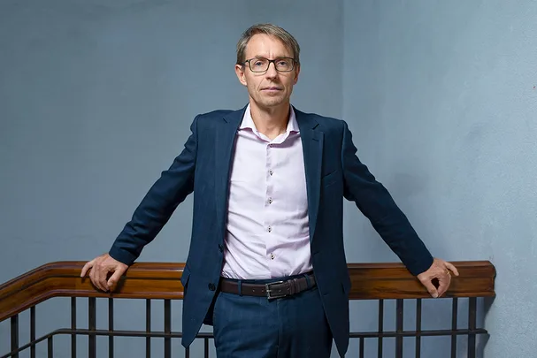 Man in a suit with glasses standing by a wooden railing in an indoor setting.