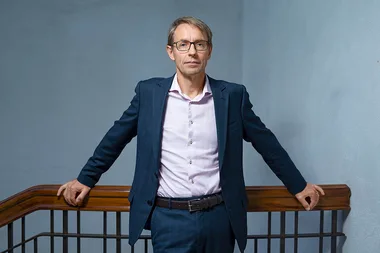 Man in a suit with glasses standing by a wooden railing in an indoor setting.