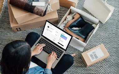 Person shopping online on a laptop surrounded by packages and shoes.
