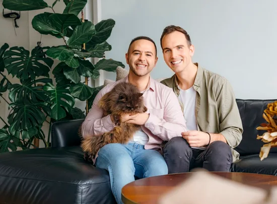 Two men smiling on a couch with a fluffy cat, surrounded by indoor plants.