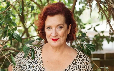 A woman with red hair and a leopard print top stands in front of green foliage, smiling at the camera.
