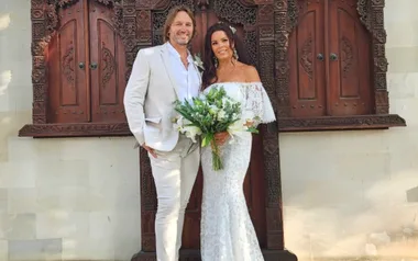 A couple in wedding attire, standing and smiling, with a decorative wooden background.