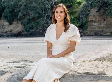Woman in a white dress sitting on a rock at the beach, with trees and a cliff in the background.