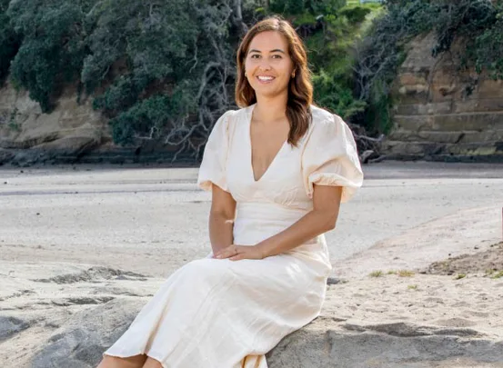 Woman in a white dress sitting on a rock at the beach, with trees and a cliff in the background.