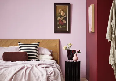 Bedroom with light pink walls, wooden headboard, striped pillow, floral artwork, and vases on a black side table.