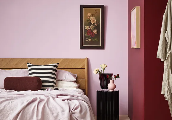 Bedroom with light pink walls, wooden headboard, striped pillow, floral artwork, and vases on a black side table.
