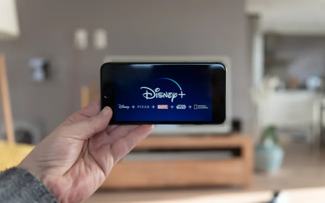 A hand holding a smartphone displaying the Disney+ logo in a living room.