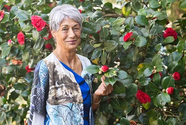 An elderly woman with short gray hair stands among bushes with red flowers, holding a branch, smiling at the camera.