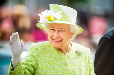 An elderly woman in a green coat and hat adorned with flowers waves and smiles warmly.