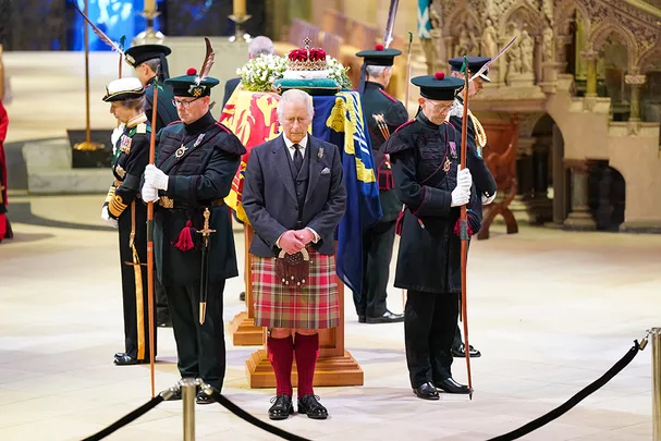 A man in a dark suit and tartan kilt stands solemnly beside a flower-covered, draped coffin, guarded by uniformed officials.