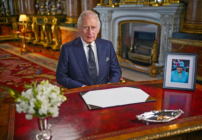 Person seated at a desk with photo of a woman in blue behind; elegant room with flowers and ornate fireplace.