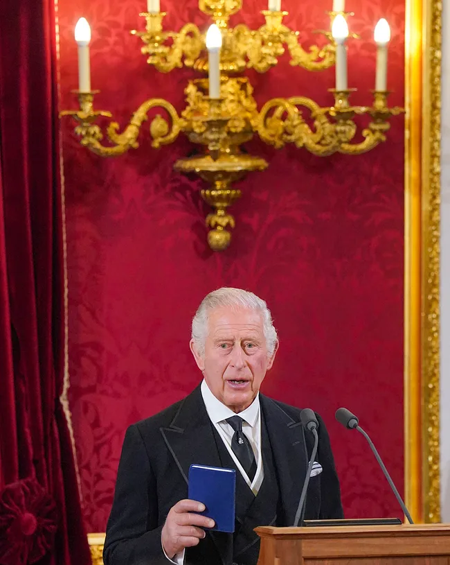 A man in a suit holds a document and speaks at a podium against an ornate red and gold background with candles.