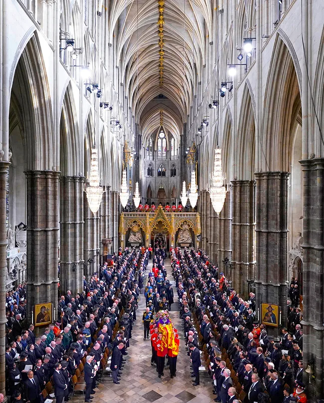 Ceremonial procession with a draped coffin in a grand cathedral, surrounded by an audience and ornate architecture.