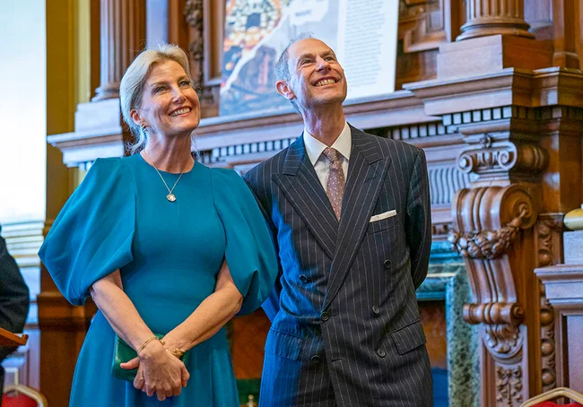 A smiling man and woman stand in a formal room, with intricate woodwork and artwork in the background.