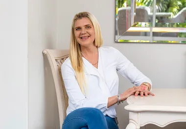 A woman in a white blouse and jeans sits smiling at a table, in a room with a window reflecting greenery outside.