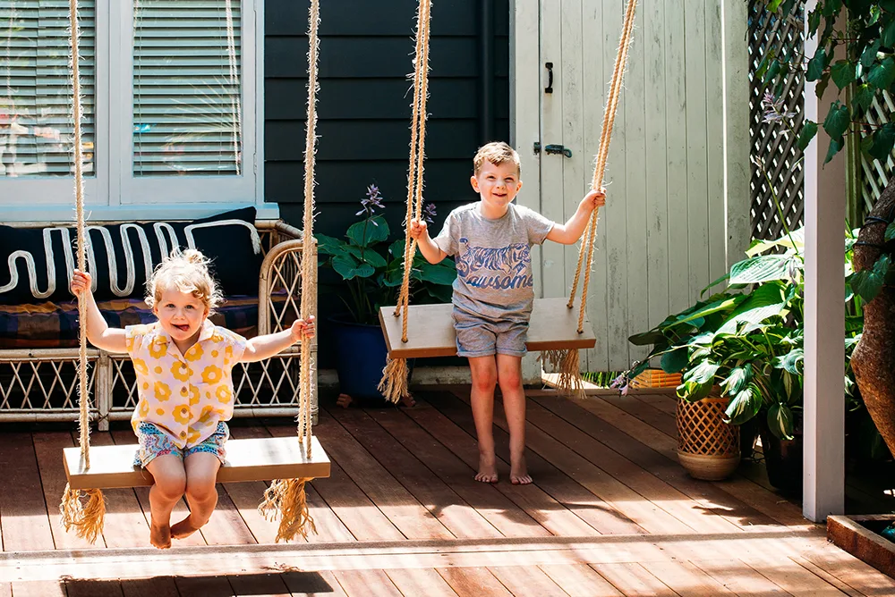 Two children play on wooden swings in a sunny backyard, surrounded by plants and outdoor furniture.