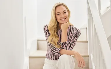 Blonde woman in a patterned blouse sitting on a staircase, smiling.