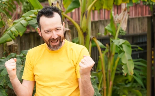 Man in a yellow shirt cheering outdoors with lush green plants in the background.