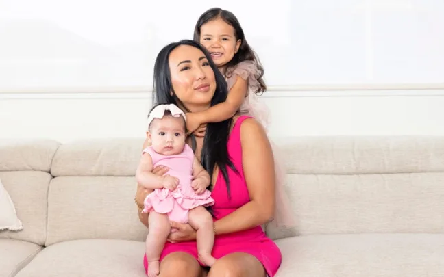 A woman in a pink dress sits on a couch holding a baby and a young girl in her arms, both in pink outfits.
