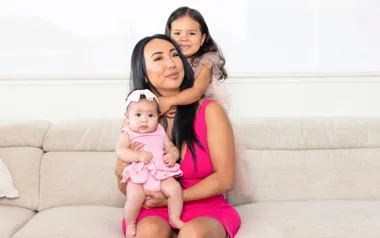 A woman in a pink dress sits on a couch holding a baby and a young girl in her arms, both in pink outfits.