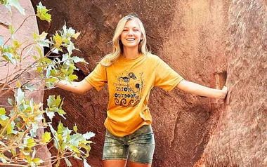 A woman smiling and standing in a canyon, wearing a yellow "Zion Outdoors" T-shirt and shorts, surrounded by rocks.