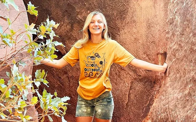 A woman smiling and standing in a canyon, wearing a yellow "Zion Outdoors" T-shirt and shorts, surrounded by rocks.
