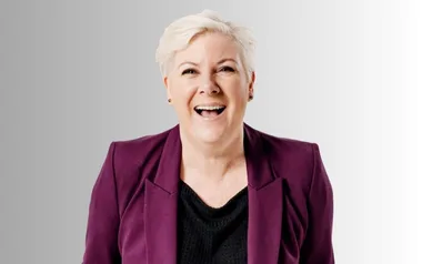Smiling person with short blonde hair in a purple blazer on a grey background.