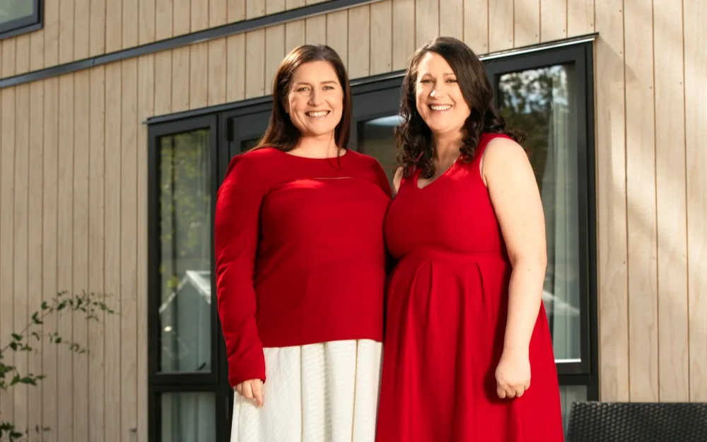 Two women in red clothing stand smiling outside by a wooden building with windows.