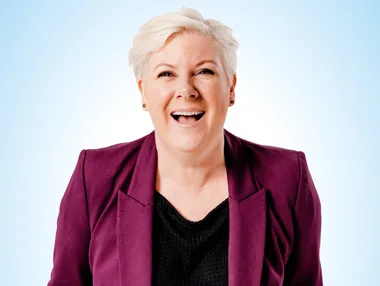 A person with short blonde hair in a purple blazer and black shirt, smiling against a light blue background.