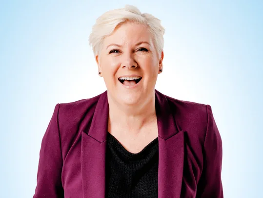 A person with short blonde hair in a purple blazer and black shirt, smiling against a light blue background.