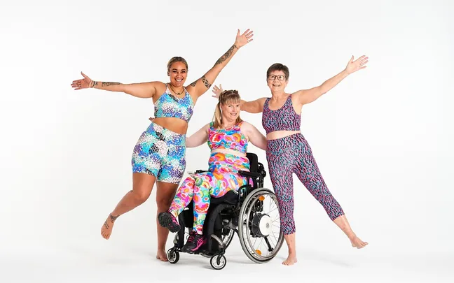 Three women in colorful activewear, one in a wheelchair, pose joyfully with arms outstretched against a white background.