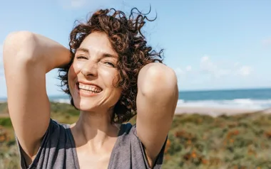 A smiling woman with curly hair stands outdoors by the beach, raising her arms behind her head on a sunny day.