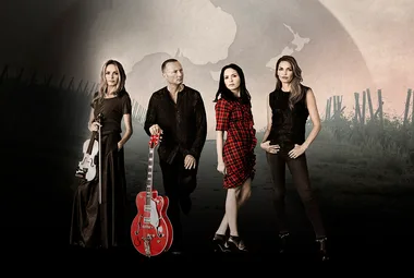 Four people stand in rural setting; one holds a violin, another a red guitar; one wears a red checkered dress.